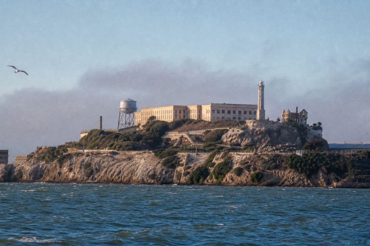 Scaffolding and construction netting along one wing of the Alcatraz cell-house exterior, the rest of the historic building open and weathered above the bare rock.