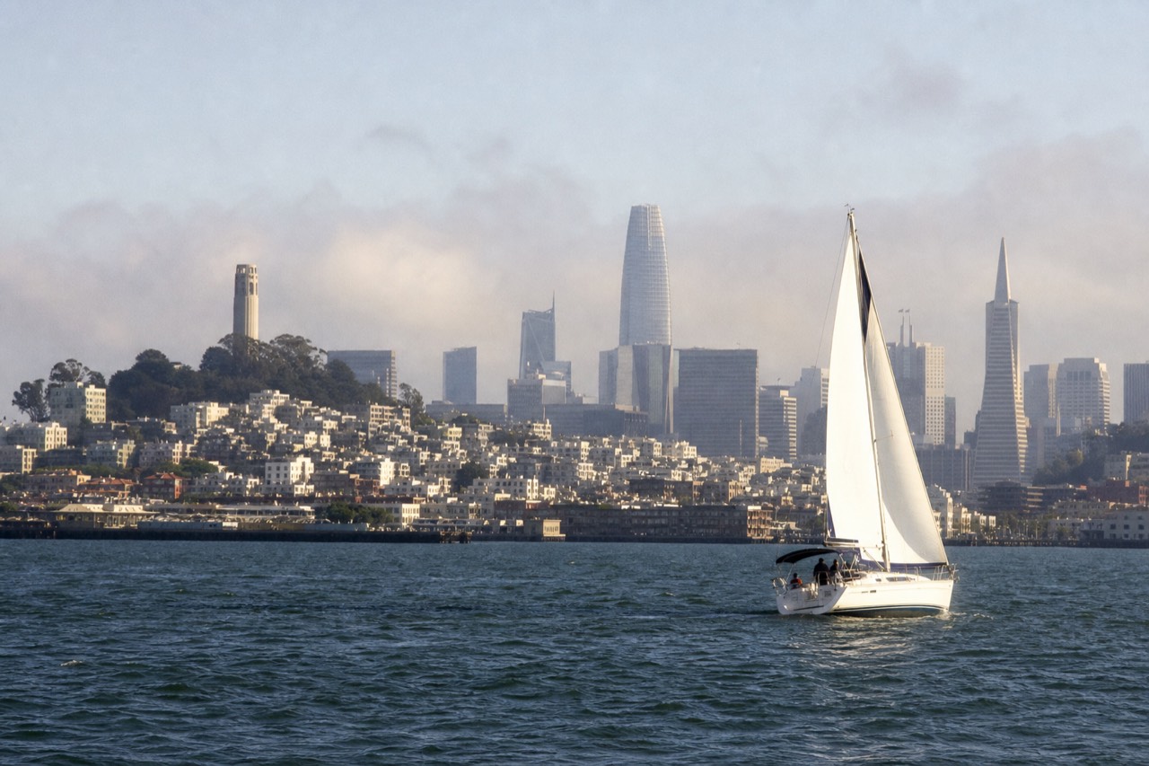 A sightseeing boat turning below the Golden Gate Bridge in warm late light, the water gold and choppy, green headlands rising into thin fog behind the towers.