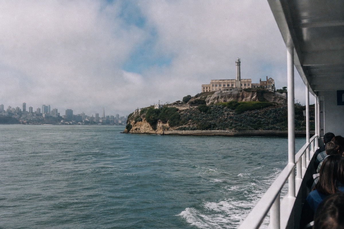 A bay cruise boat passing close to Alcatraz Island, the cellhouse and lighthouse on the rock above the water, the city skyline in the distance