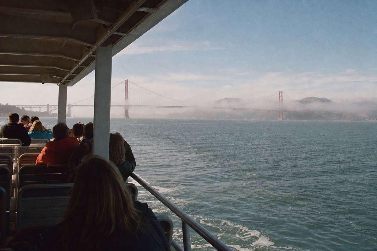 A covered sightseeing cruise boat on San Francisco Bay heading toward the Golden Gate Bridge, passengers on the open upper deck under a clear sky