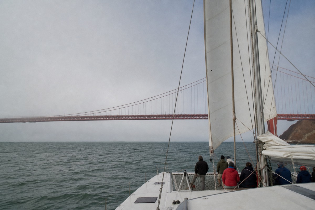 A sailing catamaran under full sail on San Francisco Bay, a small group of guests aboard, the Golden Gate Bridge low behind the rigging