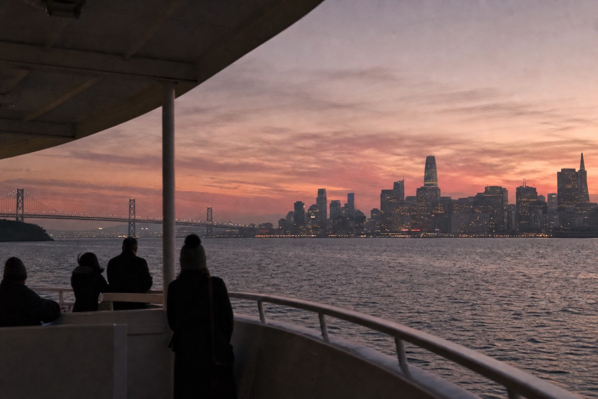 A bay cruise boat at dusk, the San Francisco skyline and the bridges lighting up against a pink and gold sky over the water