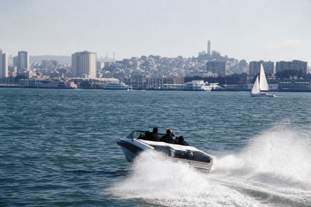 A high-speed jet boat throwing up spray as it turns hard on San Francisco Bay, the city waterfront behind