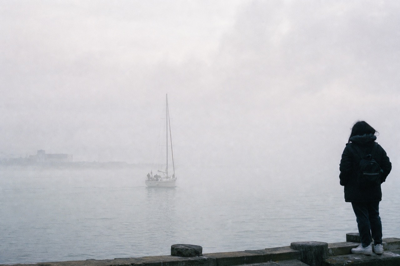 A white sightseeing boat passing beneath the south tower of the Golden Gate Bridge on grey-green water, the rust-red steel rising straight overhead into thin fog.
