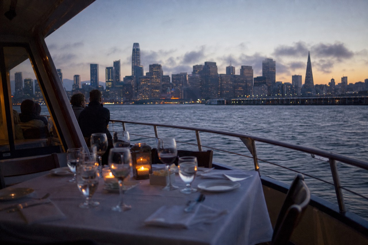 A window table set with white linen and glassware aboard a boat at night, blurred city lights glowing beyond the dark glass.