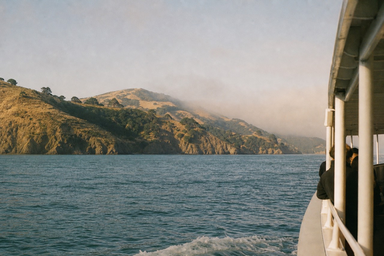 A couple in warm jackets at a boat rail at dusk, wind in their hair, the darkening bay and a distant lit skyline stretching behind them.